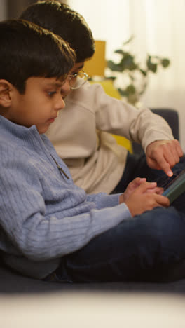 Video-Vertical-De-Dos-Niños-Pequeños-Sentados-En-Un-Sofá-En-Casa-Jugando-O-Transmitiendo-Juntos-A-Una-Tableta-Digital